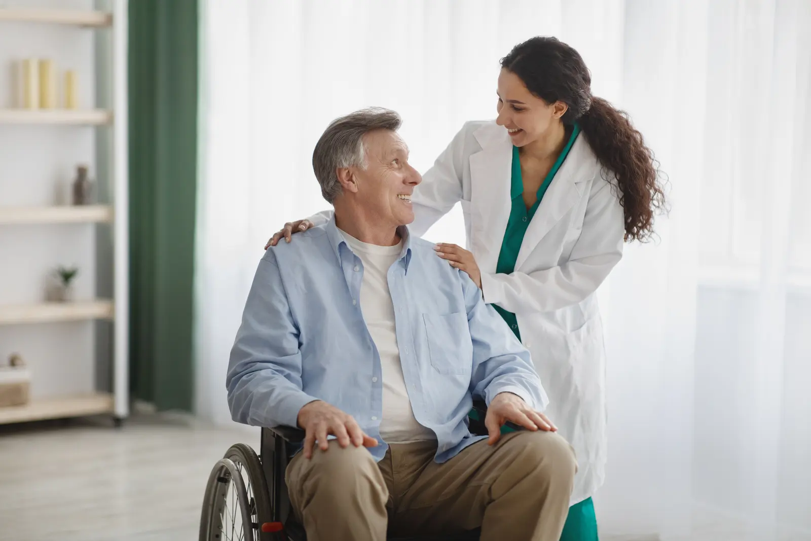 An older woman in a wheelchair being wheeled slowly through a sunlit room
